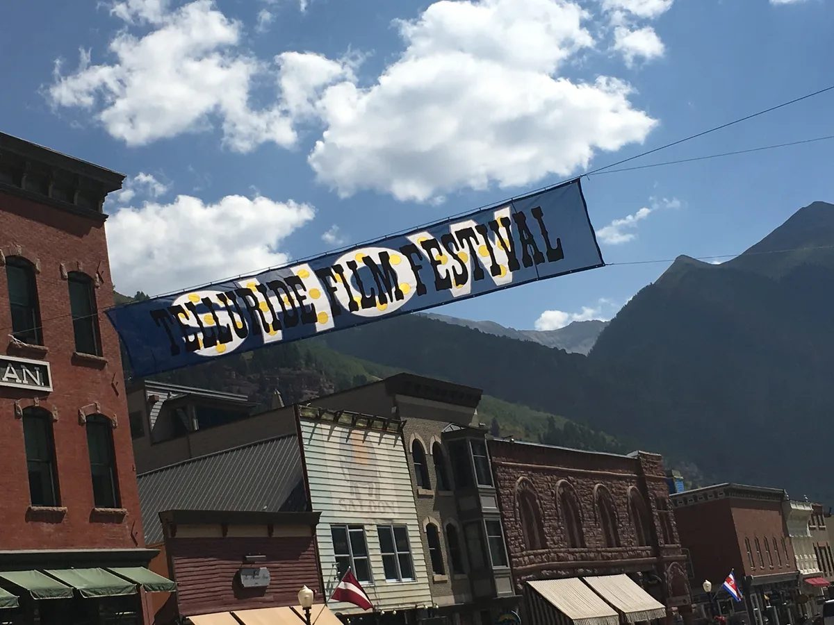 Colorado Avenue in Telluride is lined with historic brick and wood storefronts beneath a bright blue sky, while a large “Telluride Film Festival” banner stretches overhead. Steep dark mountain walls rise behind the town, framing the street in the narrow box canyon.