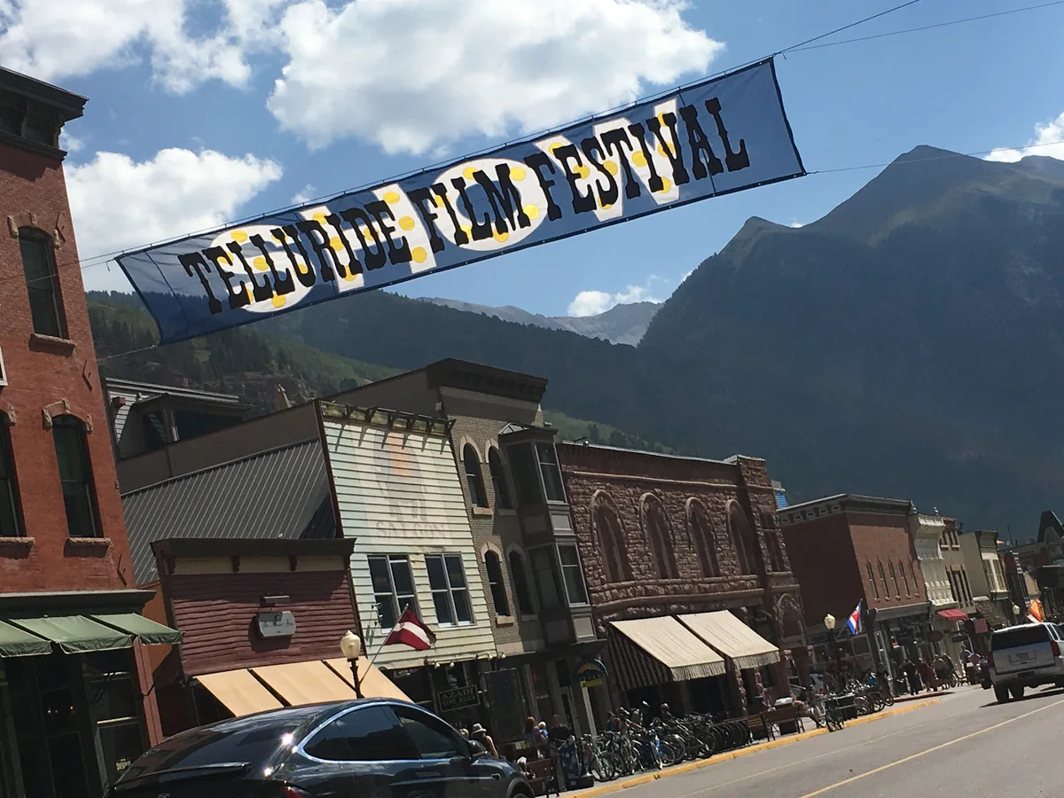 Colorado Avenue in Telluride is lined with historic brick and wood storefronts beneath a banner reading “Telluride Film Festival,” with pedestrians, parked bikes, and cars along the street. Steep, dark mountain walls rise sharply behind the town under a bright blue sky with scattered clouds.