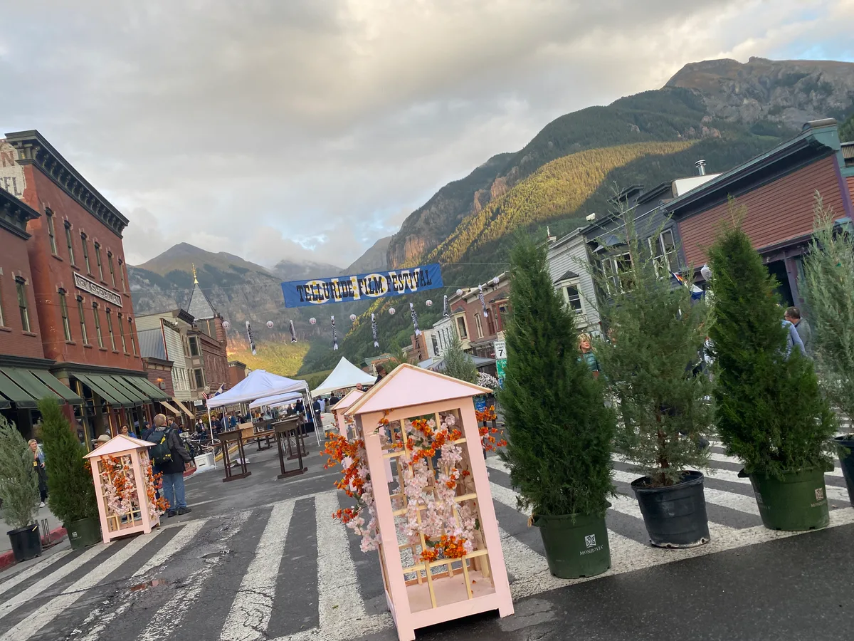 Colorado Avenue in Telluride is closed for the film festival, with white tents, pink floral display stands, and potted evergreen trees lined up along the street. Victorian brick storefronts and a “Telluride Film Festival” banner lead the eye toward steep mountain walls glowing in soft late-day light.