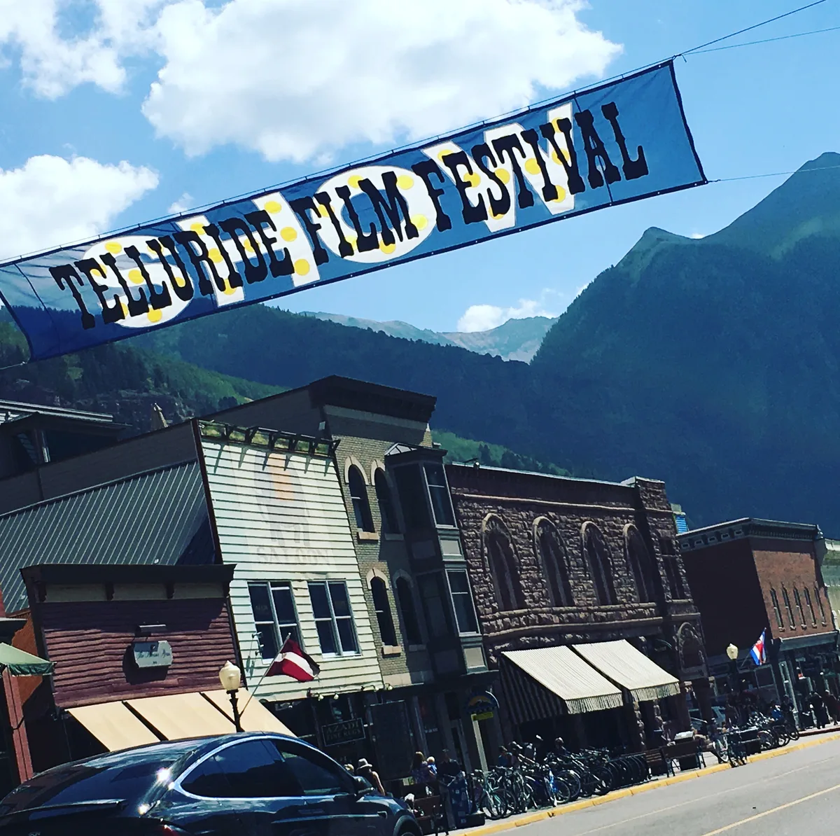 A diagonal blue banner reading “Telluride Film Festival” stretches across Colorado Avenue above Victorian storefronts, with parked bikes and cars lining the street below. Steep green mountain slopes rise behind the town under a bright blue sky with scattered clouds.
