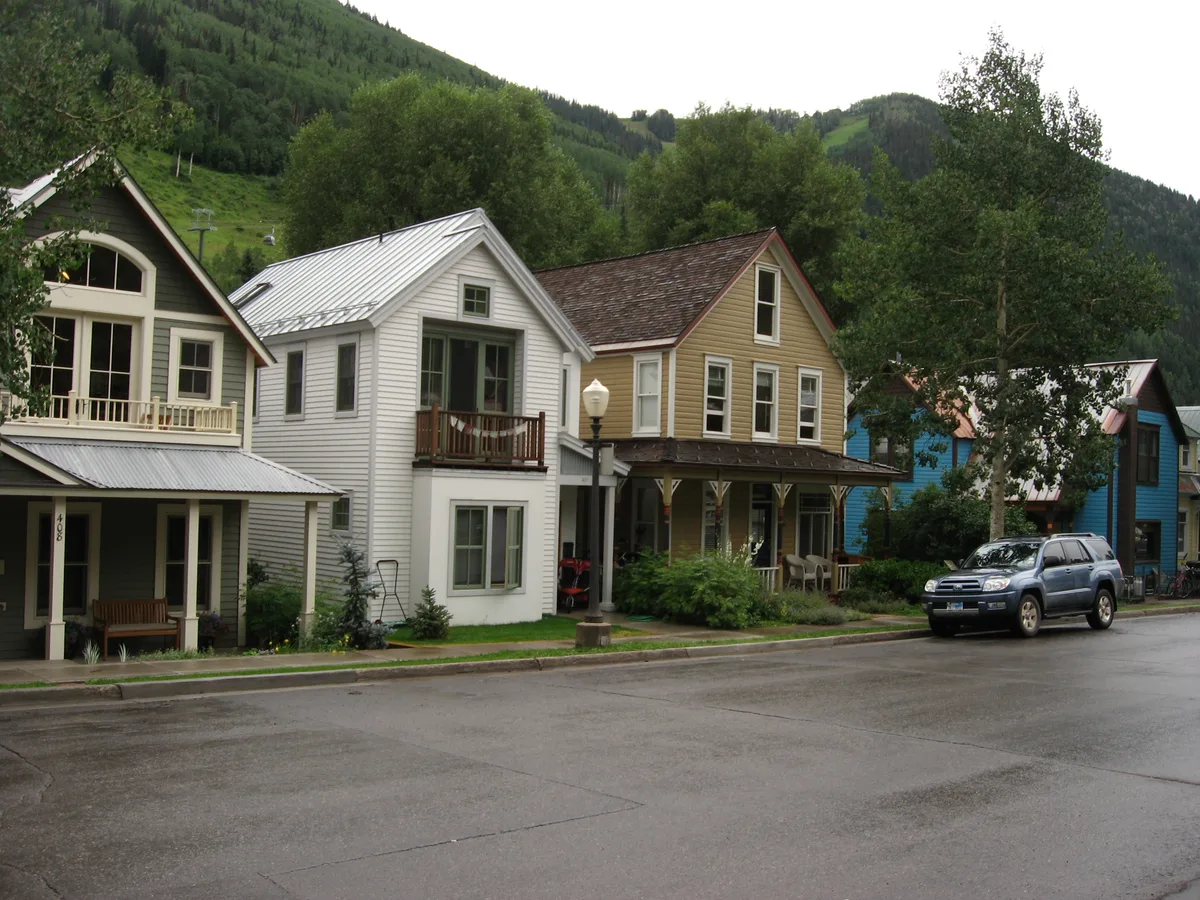 Colorful Victorian-style houses line a quiet street in Telluride, with porches, gabled roofs, and a parked SUV in the foreground. Steep green mountain slopes rise behind the homes under a pale sky.