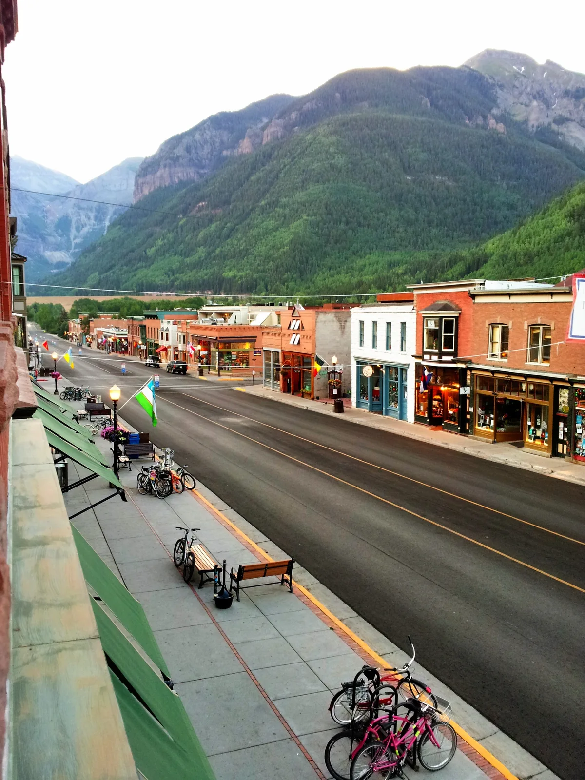 Colorado Avenue runs straight through Telluride’s Victorian downtown, with colorful storefronts, parked bikes, and flags lining the quiet street. Steep green mountain walls rise dramatically behind the town, filling the box canyon under a bright, hazy sky.