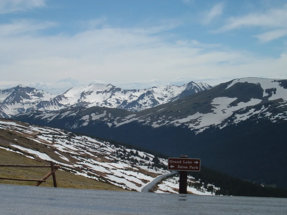 Snow-speckled alpine ridges stretch across the horizon under a pale blue morning sky, with the dark Forest Canyon dropping away below. In the foreground, a Trail Ridge Road overlook sign points toward Grand Lake and Estes Park beside a curving road and patchy tundra slopes.