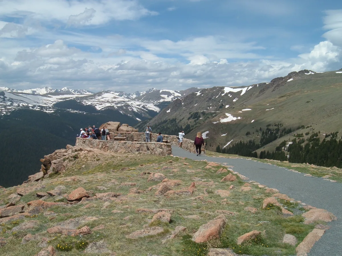 Visitors stand on a stone overlook beside a paved path above a high alpine slope, with rocky tundra and a few tiny wildflowers in the foreground. Beyond the railing, snow-streaked mountains and a deep forested canyon stretch under a partly cloudy blue morning sky.