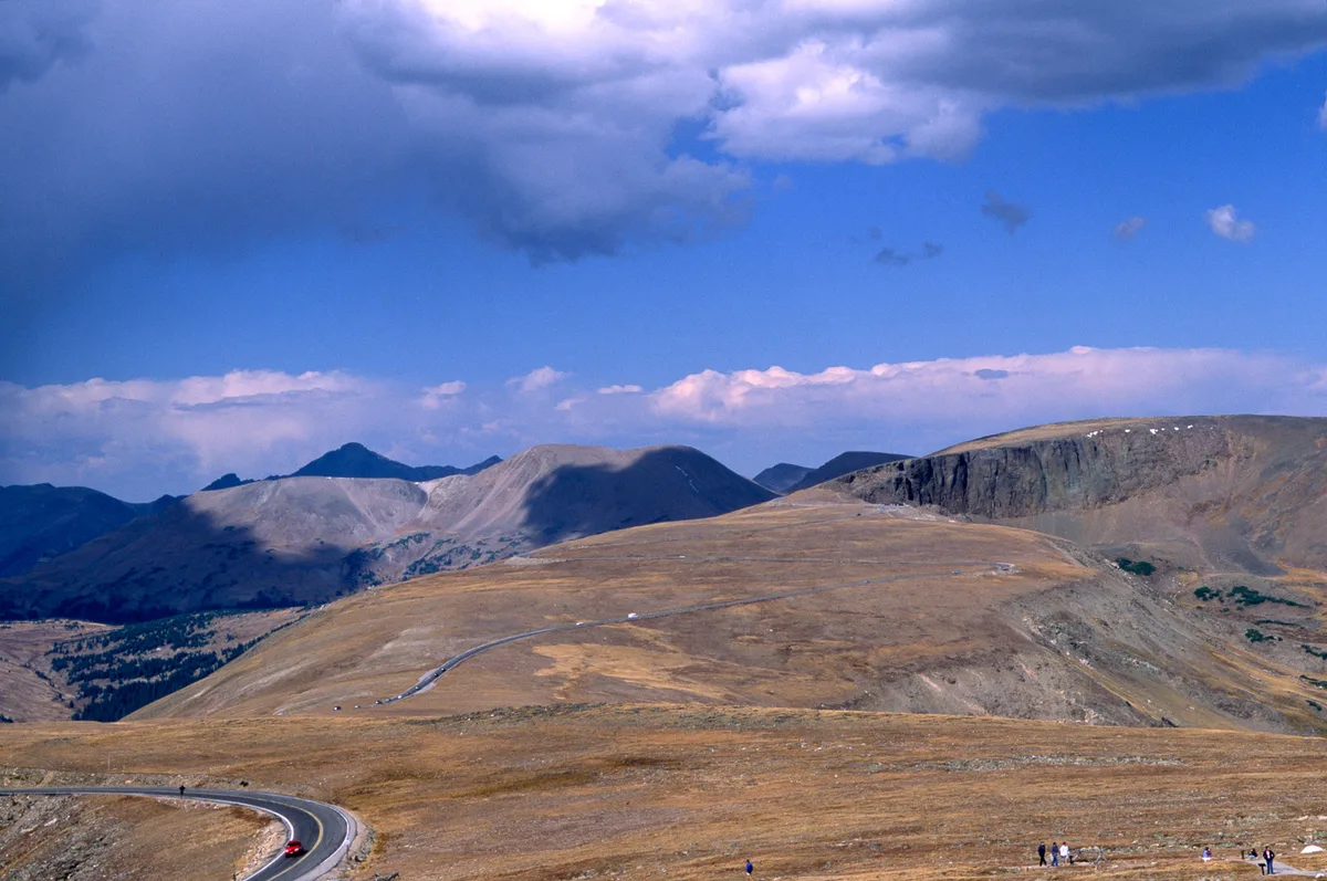 Trail Ridge Road curves through golden alpine tundra in the foreground, with a red car and a few tiny visitors emphasizing the vast scale of the landscape. Beyond, rounded high peaks and the steep dark wall of Forest Canyon sit under a dramatic blue sky with heavy clouds.