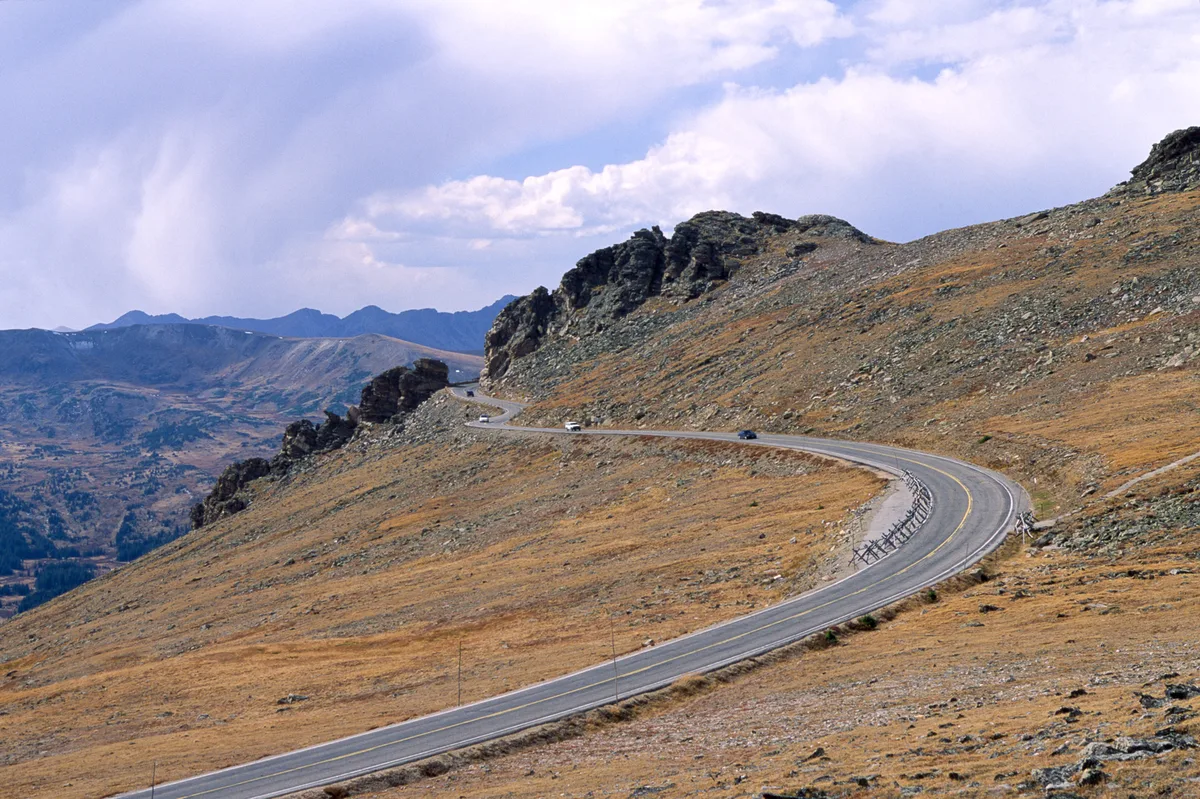 Trail Ridge Road curves in a long S across the barren alpine tundra, with a few cars tracing the pavement beneath dark rocky outcrops. Distant mountain ridges and a broad, cloud-filled sky open up the view across the Continental Divide.