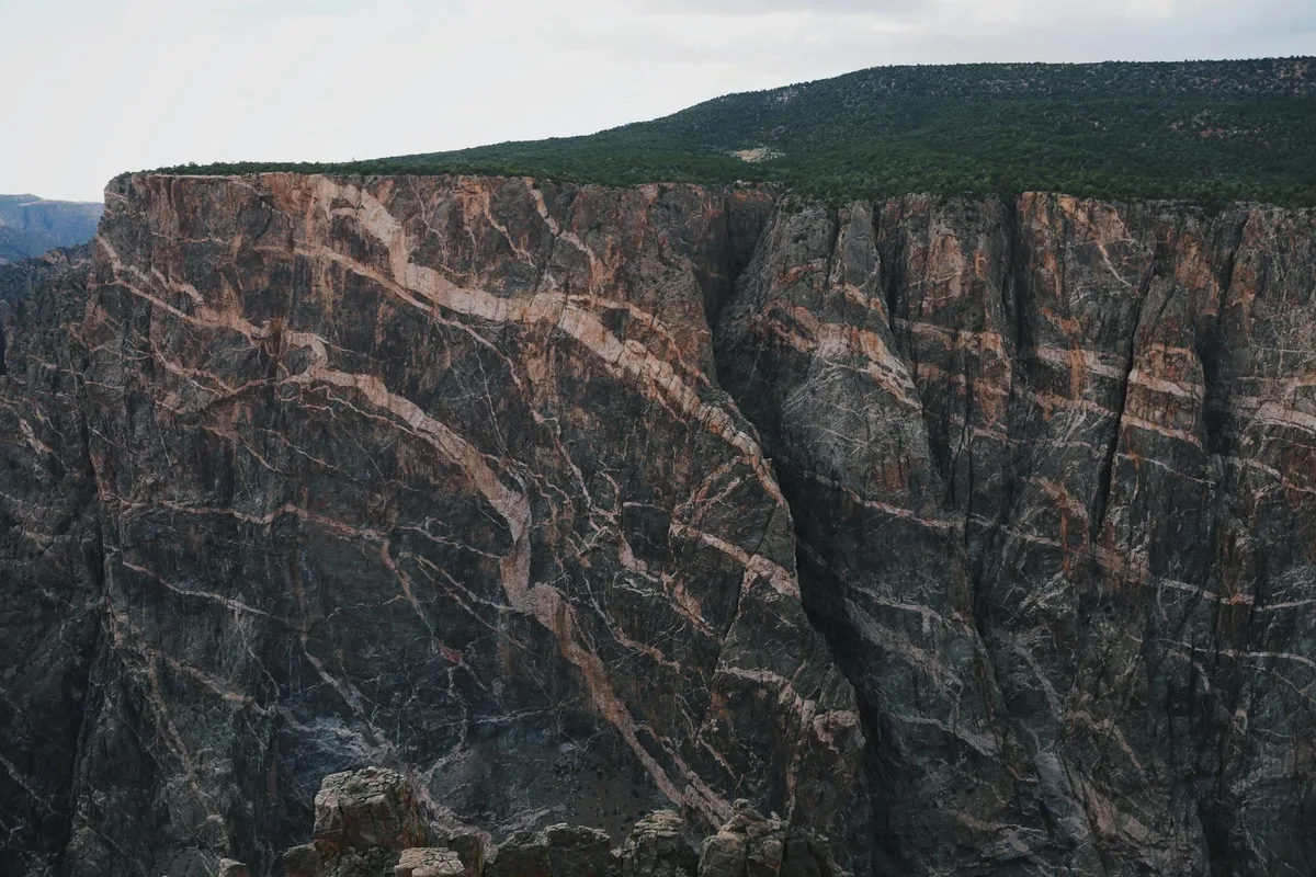 A sheer, striped cliff wall drops into a deep canyon, with a dark green tundra ridge lining the top against a pale morning sky. The rock face shows dramatic vertical seams and diagonal mineral bands, emphasizing the scale and steepness of the overlook.