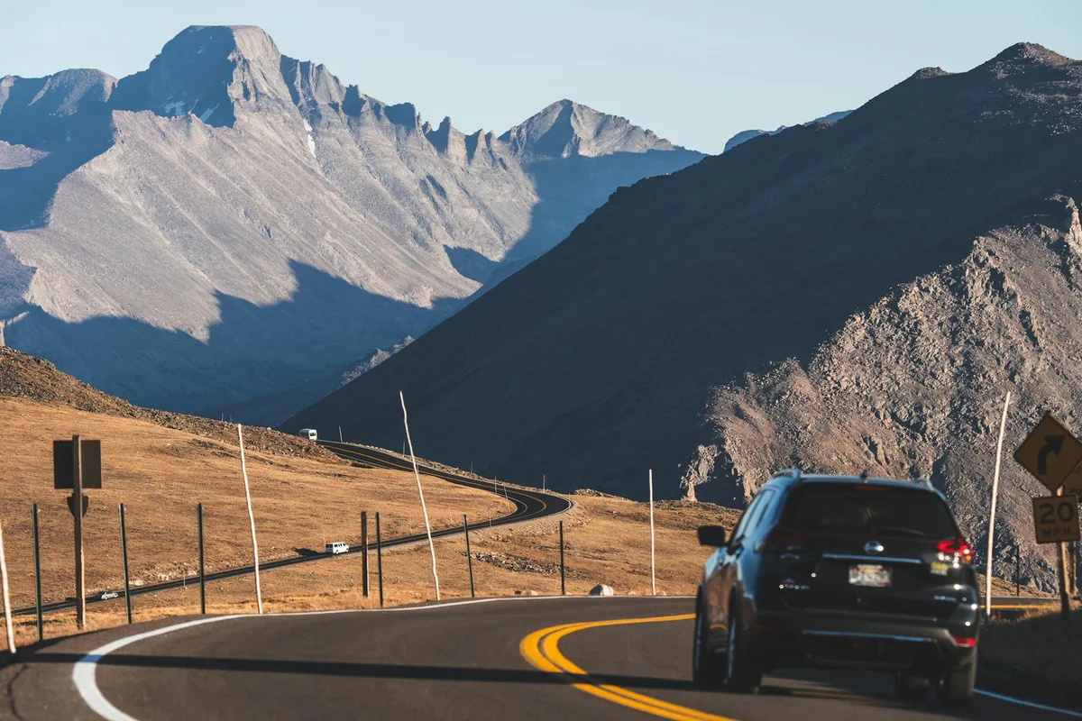 A winding alpine highway curves through tawny tundra beneath steep, shadowed mountain walls, with a black SUV in the foreground and a small vehicle farther ahead. Bright morning light catches the gray peaks and rocky ridges of the Continental Divide under a clear blue sky.