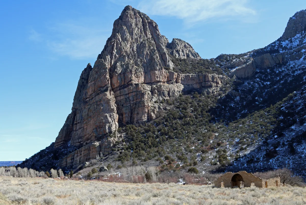 A towering gray-and-rust sandstone spire rises above a sunlit canyon slope dusted with patchy snow and dark evergreen shrubs. In the lower right, a small stone ruin sits in a grassy meadow beneath the sheer rock wall under a clear blue afternoon sky.