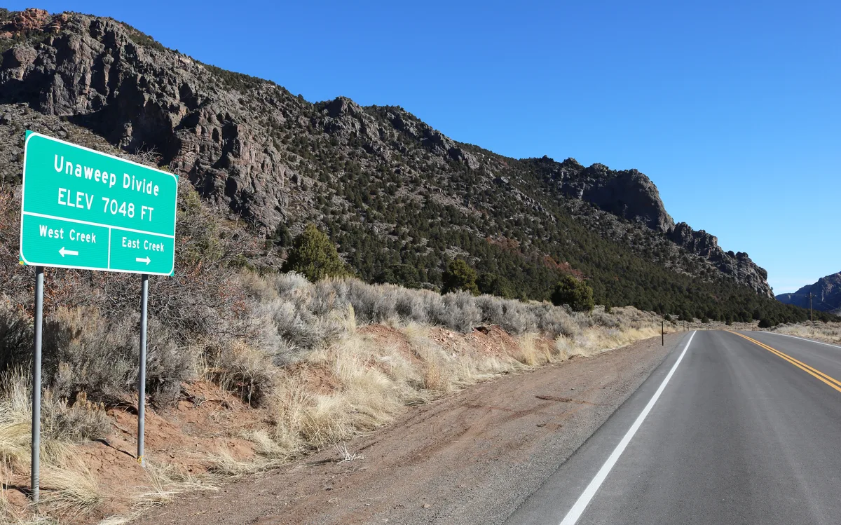 A green roadside sign marks the Unaweep Divide at 7,048 feet, with arrows pointing west to West Creek and east to East Creek. Highway 141 curves into a wide, sunlit canyon of rugged dark rock walls, sparse shrubs, and a clear blue sky.