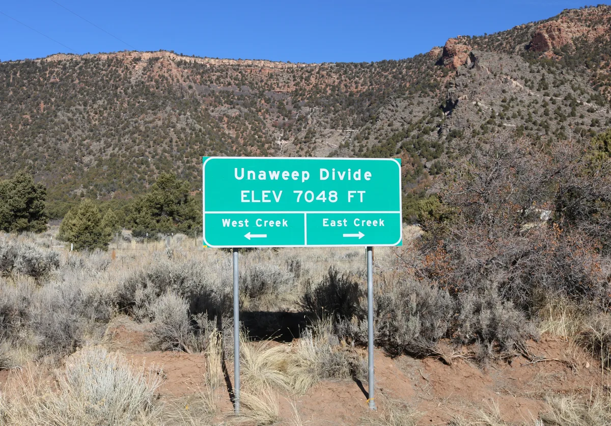 A green roadside sign for “Unaweep Divide” stands in the foreground, marking elevation 7,048 feet and pointing left to West Creek and right to East Creek. Behind it, sunlit canyon walls rise under a clear blue sky, with sparse shrubs and dry grass in the foreground.