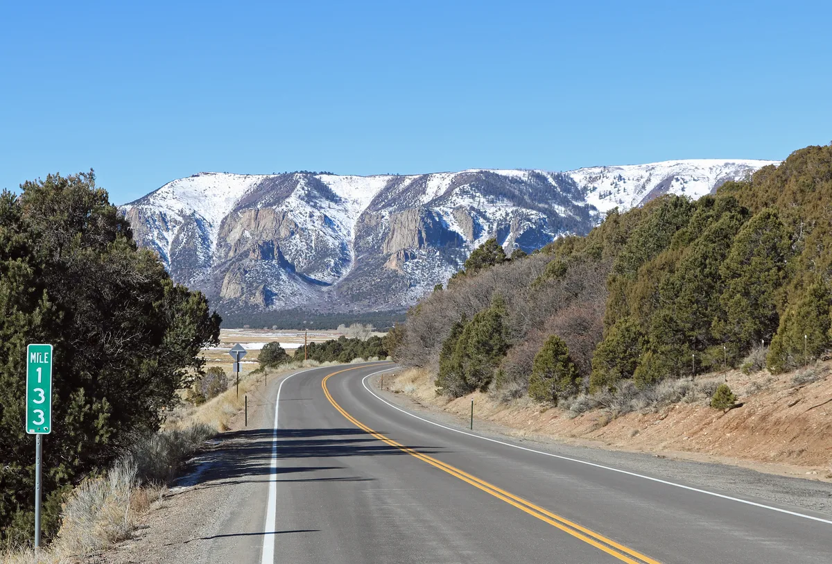 A winding two-lane road leads through Unaweep Canyon toward snow-streaked granite cliffs under a clear blue sky. Evergreen-covered slopes frame the scene, with a mile marker and roadside sign in the foreground.
