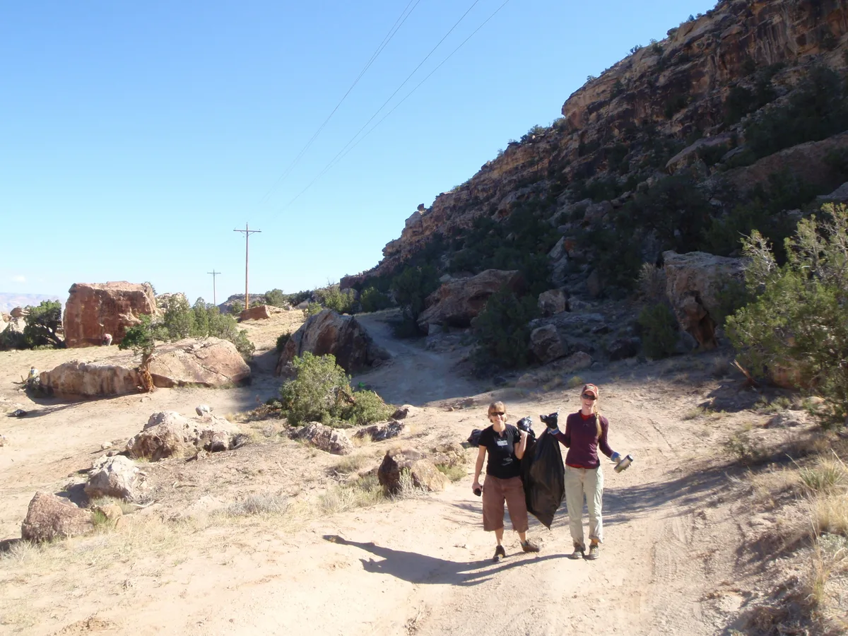 Two hikers walk along a dusty trail in Unaweep Canyon, with sunlit granite boulders and a steep, shadowed rock wall rising on the right. Power lines cut across the clear blue sky above the sparse shrubs and open canyon floor.