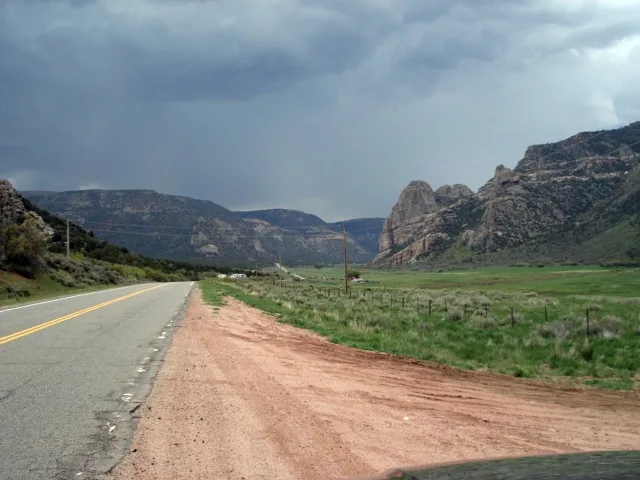 A two-lane road curves into a broad green canyon floor, with steep dark granite cliffs rising on both sides and a layered rock outcrop on the right. Heavy gray storm clouds fill the sky, giving the afternoon scene a moody, rain-soaked look.