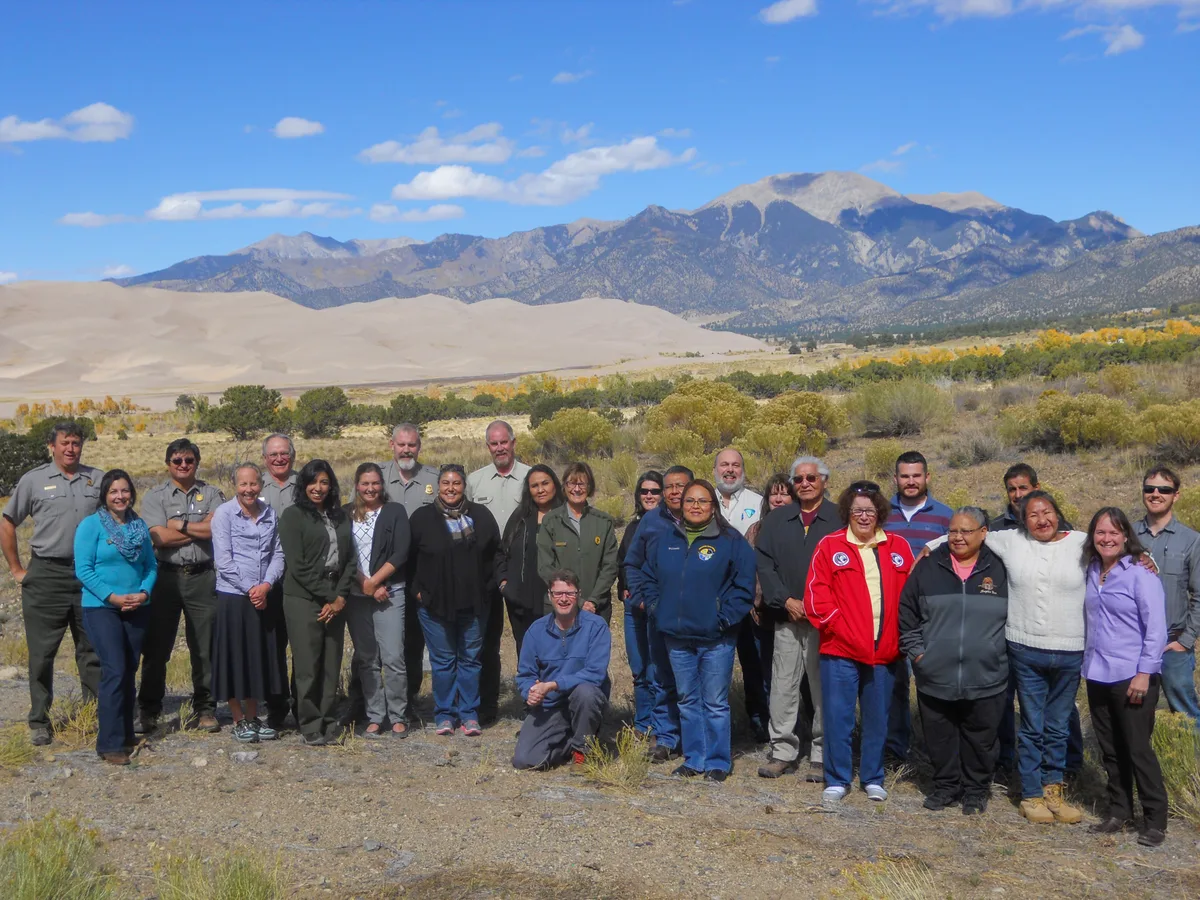 A large group of Ute Mountain Ute tribal members and NPS staff stands for a posed portrait in a scrubby high-desert landscape, with pale sand dunes and rugged mountains rising behind them under a bright blue morning sky. The scene is open and sunlit, with scattered clouds and autumn-yellow cottonwoods lining the valley below.