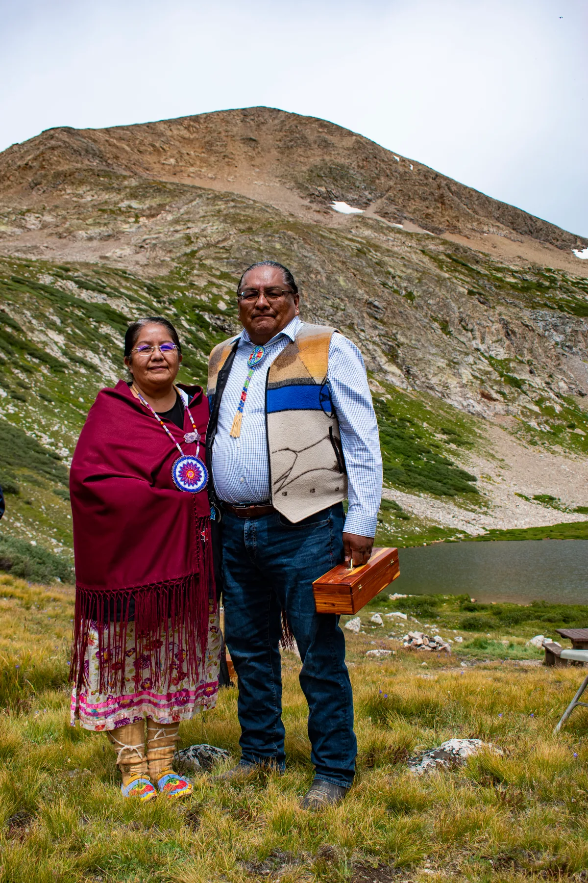 Two Ute Mountain Ute tribal members stand posed in the foreground on grassy alpine ground, wearing traditional regalia and holding a small wooden box. Behind them, a broad rocky mountain slope rises above a small lake under an overcast morning sky.