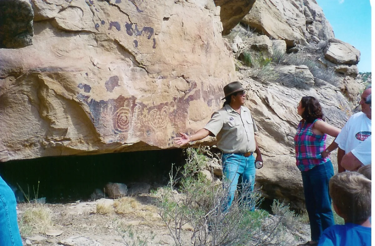 A Ute Mountain Ute guide stands beside a sandstone alcove covered with faded petroglyphs, explaining the site to a small group of visitors. Morning light highlights the tan rock face, dark cave opening, and sparse desert brush in the canyon.