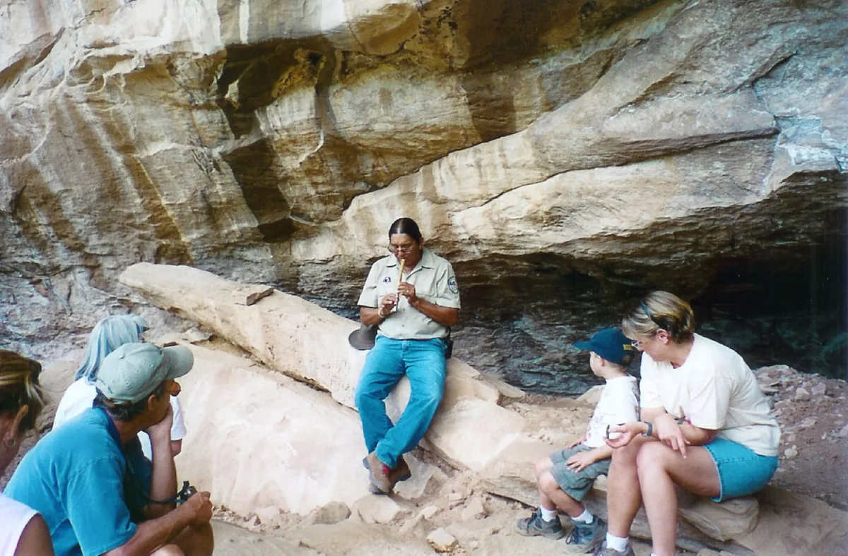 A Ute Mountain Ute guide sits on a pale rock ledge beneath a broad sandstone overhang, speaking to a small seated group of visitors in the cool shade. The canyon wall fills the background with layered tan and gray striations, lit by soft morning light.