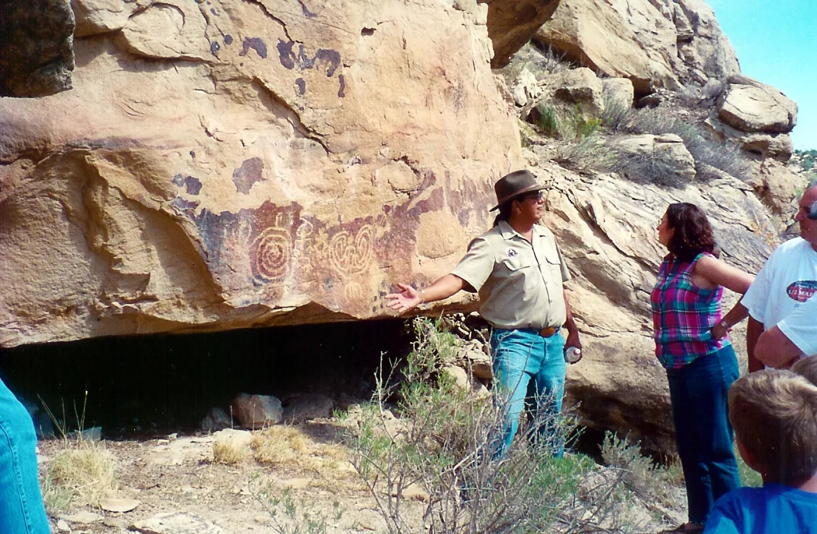 A Ute Mountain Ute guide stands beside a sunlit sandstone ledge covered with faded petroglyphs, speaking to a small group gathered at the mouth of a shallow rock shelter. The scene shows a dry, remote canyon landscape with scrubby plants, rough boulders, and clear morning light.