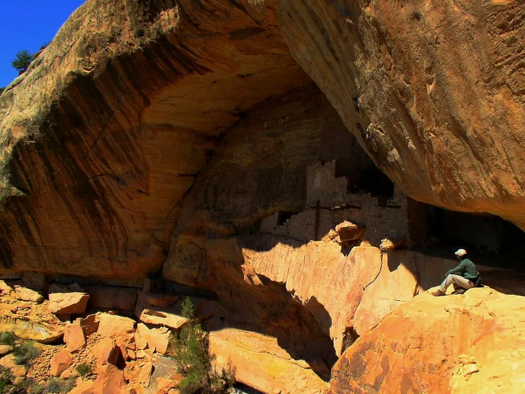 A massive sandstone alcove shelters the remains of an Ancestral Puebloan cliff dwelling, with stacked stone walls tucked deep in shadow beneath the overhang. Sunlit orange rock ledges fill the foreground, and a lone seated visitor on the right gives a sense of the site’s scale.