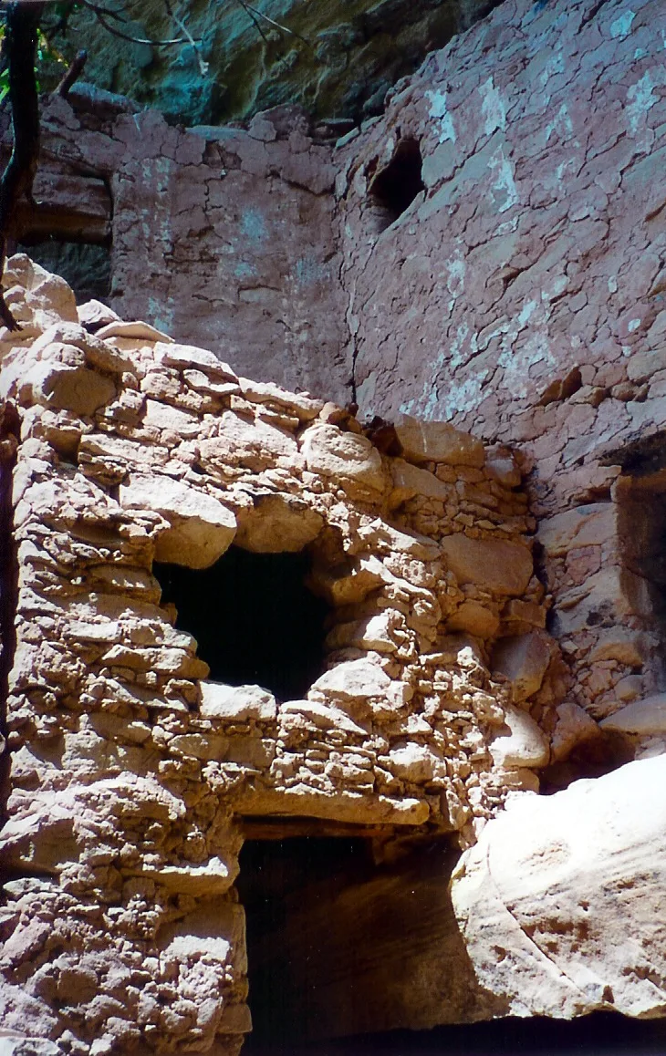 Sunlit cliff dwellings built into a narrow canyon wall, with rough stacked stone rooms and dark doorways set against a weathered pink-beige rock face. Bright morning light highlights the masonry while the upper recesses stay shaded by the overhang.