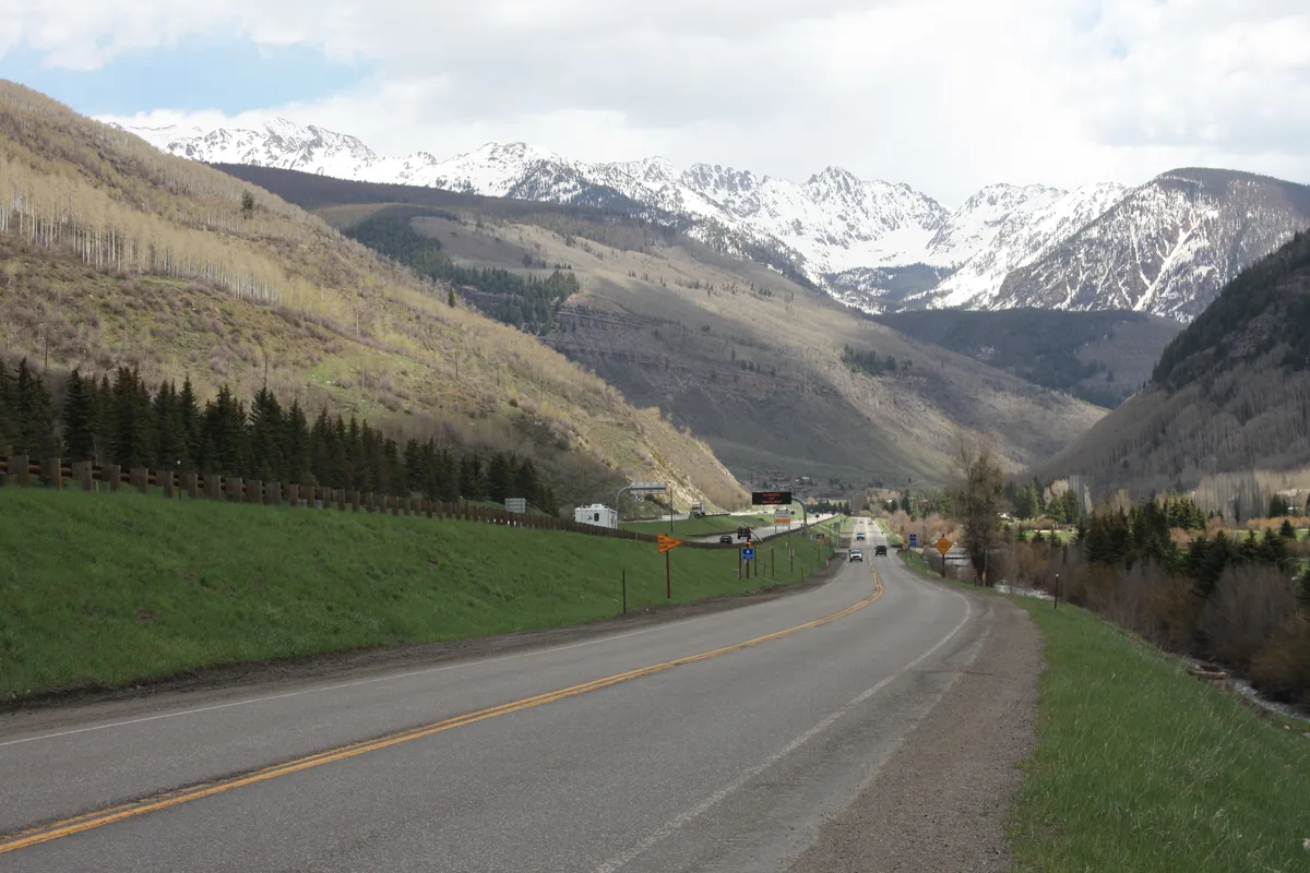 A winding road leads into a broad mountain valley beneath the snow-covered, jagged Gore Range, with dark evergreen slopes and pale spring-green hillsides on either side. Soft afternoon light and scattered clouds brighten the alpine landscape in the distance.