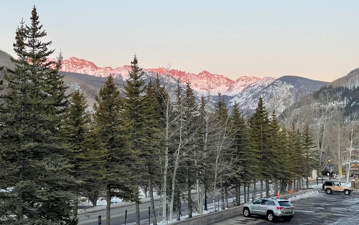 Snow-dusted Gore Range peaks glow pink in the evening light above a line of evergreen trees and leafless aspens. In the foreground, a parking area and road sit at the base of the mountain valley under a clear pale sky.