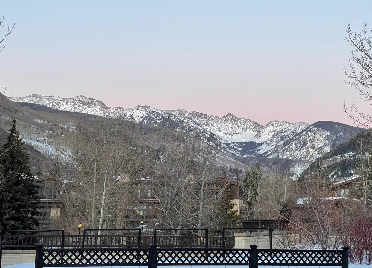 Snow-dusted Gore Range peaks stretch across the horizon under a pale pink evening sky, with dark evergreen slopes and bare winter trees in the foreground. Lodge rooftops and a black lattice fence frame the view from the overlook.
