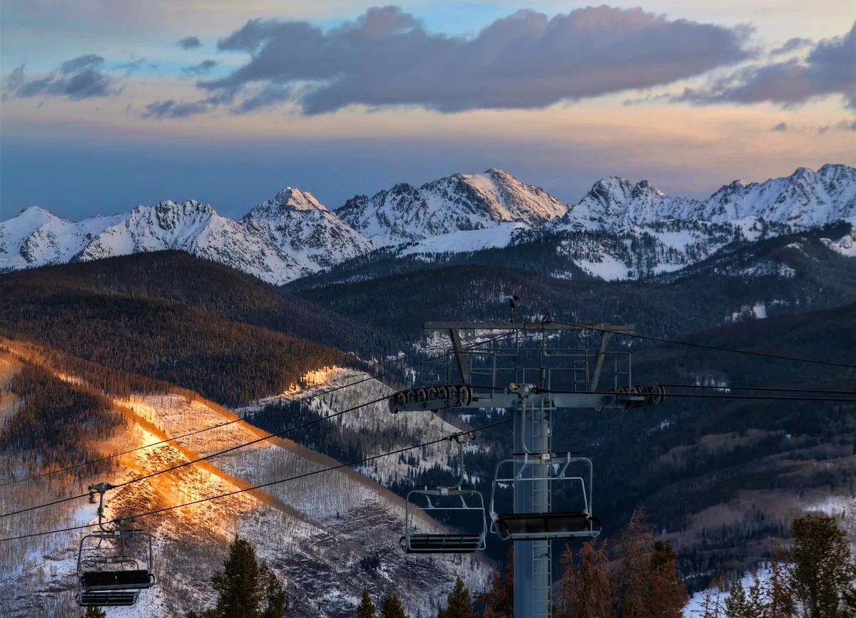 A chairlift rises in the foreground over dark, forested slopes, with a warm strip of sunlight lighting the left hillside. Snow-covered Gore Range peaks stretch across the horizon under a pastel evening sky with low clouds.