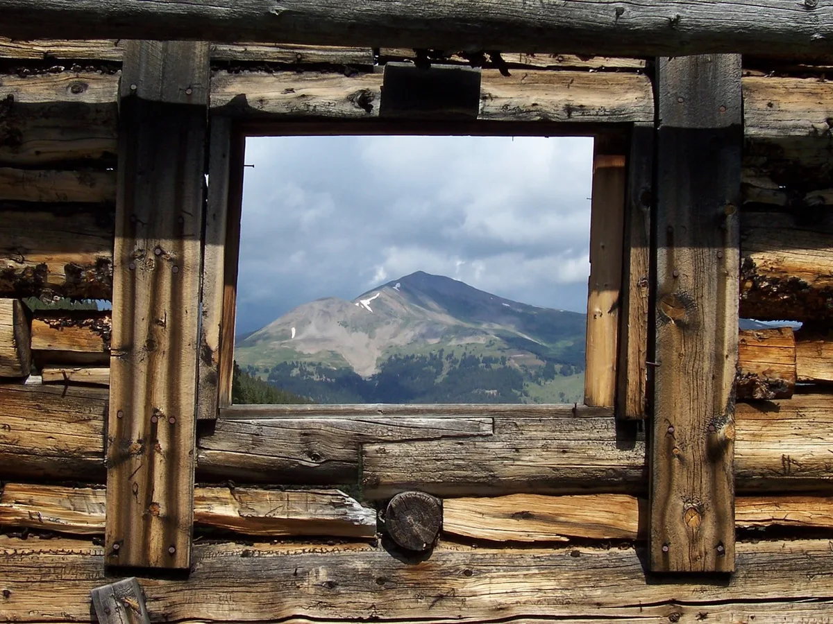 Weathered log walls and a square window frame a distant mountain peak under heavy gray clouds, creating a rustic, postcard-like view. The rough wood grain and dark support beams contrast with the pale alpine slopes and small patches of lingering snow.