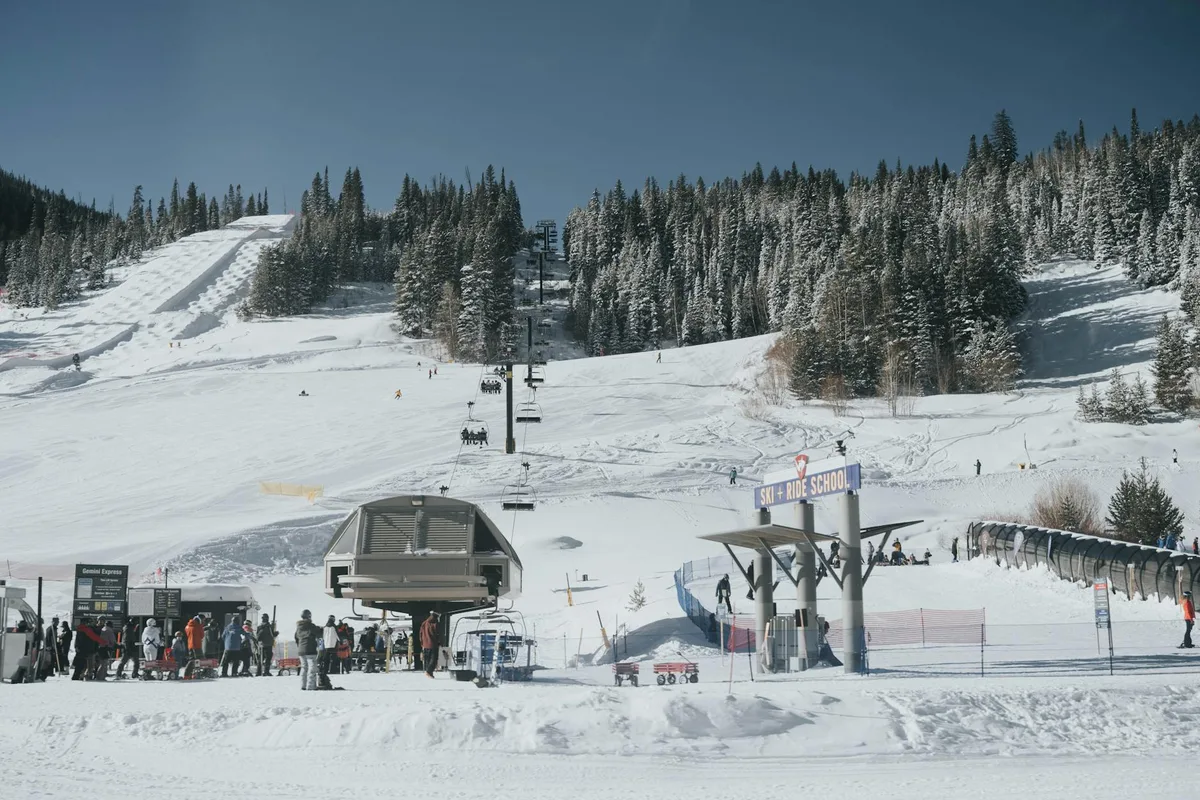 Snowy ski slopes stretch up toward a dense line of evergreen trees under a clear blue sky, with chairlifts running up the center of the mountain. In the foreground, skiers gather near the gondola station and a “Ski + Ride School” sign, with tracks etched across the bright snow.