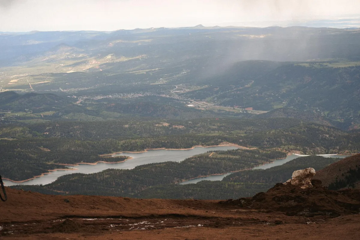 Sweeping mountain overlook with hazy afternoon light over a broad valley, dark forested ridges, and a chain of pale blue reservoirs below. A rocky dirt foreground frames the view, with distant hills fading into the bright horizon.