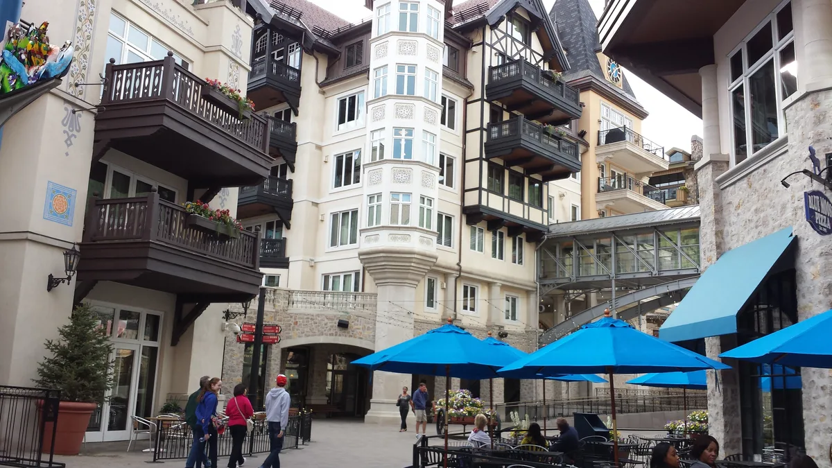 Tall cream-colored alpine buildings with dark wooden balconies frame a pedestrian courtyard, with bright blue café umbrellas and people walking below. A glass-enclosed skybridge links the buildings overhead, adding to the layered European ski-village feel.