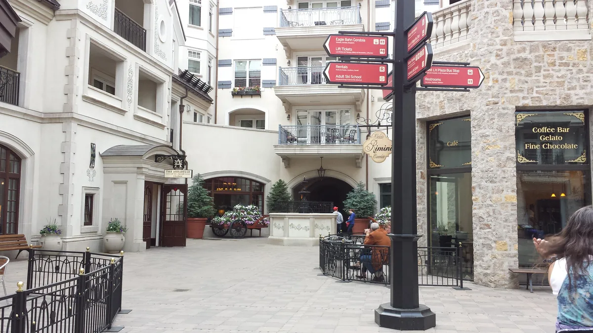 A pedestrian plaza in Vail Village with cream-colored alpine-style buildings, stone accents, balconies, and flower planters around a central courtyard. Red directional signs, café windows, and a few people seated outside add to the busy mountain-town atmosphere.