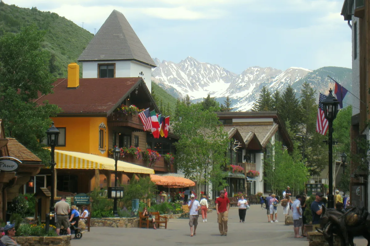 Pedestrian street in Vail Village with colorful Bavarian-style buildings, striped awnings, hanging flower baskets, and flags lining the walkway. Snowy mountain peaks rise in the background behind the tree-filled village center.