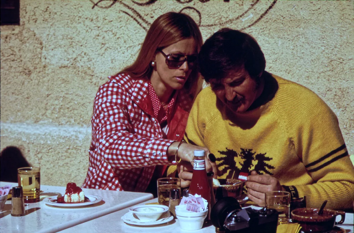 Two people sit at an outdoor café table in Vail Village, leaning over a meal with drinks, ketchup, and a camera on the table. The scene is framed tightly against a stucco wall in warm sunlight, with the woman in a red-and-white checkered jacket and the man in a bright yellow sweater.