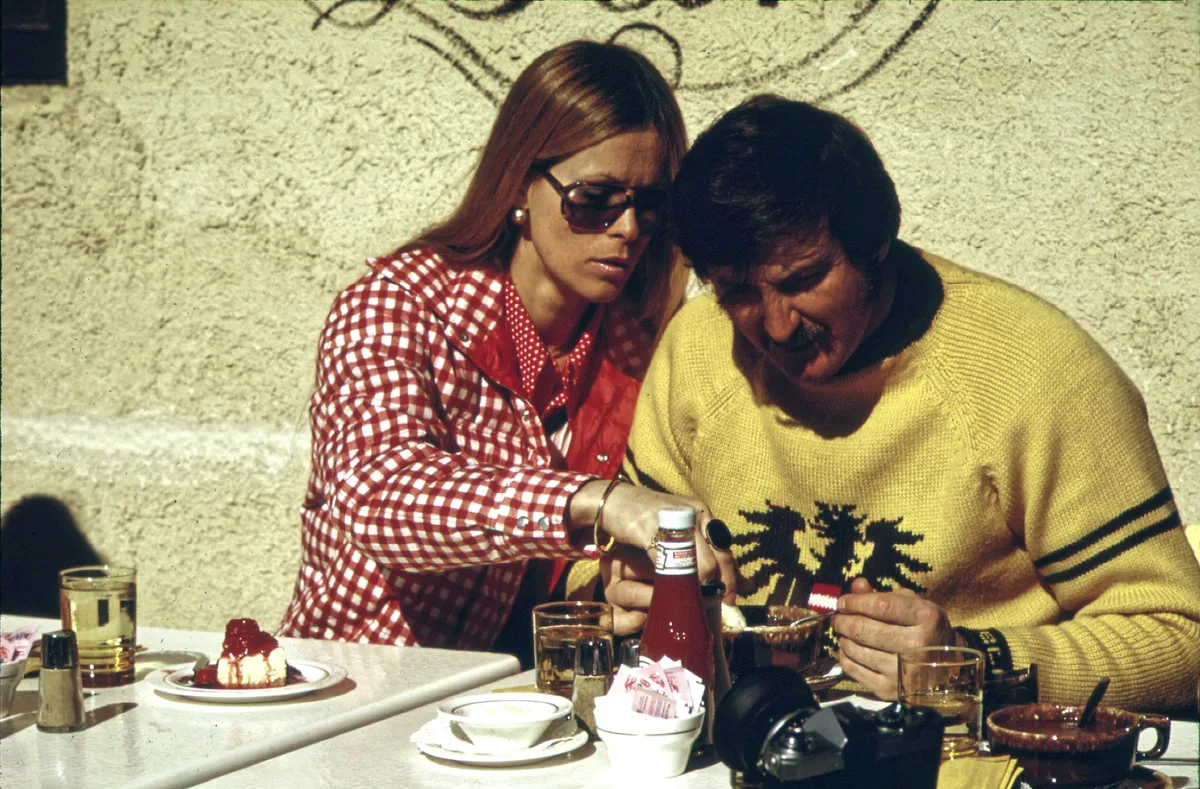 Two people sit at an outdoor café table in bright sunlight, with drinks, a dessert, and a ketchup bottle spread across the white tabletop. The woman in sunglasses leans in beside the man in a yellow sweater as they focus on the meal, with a textured stucco wall filling the background.