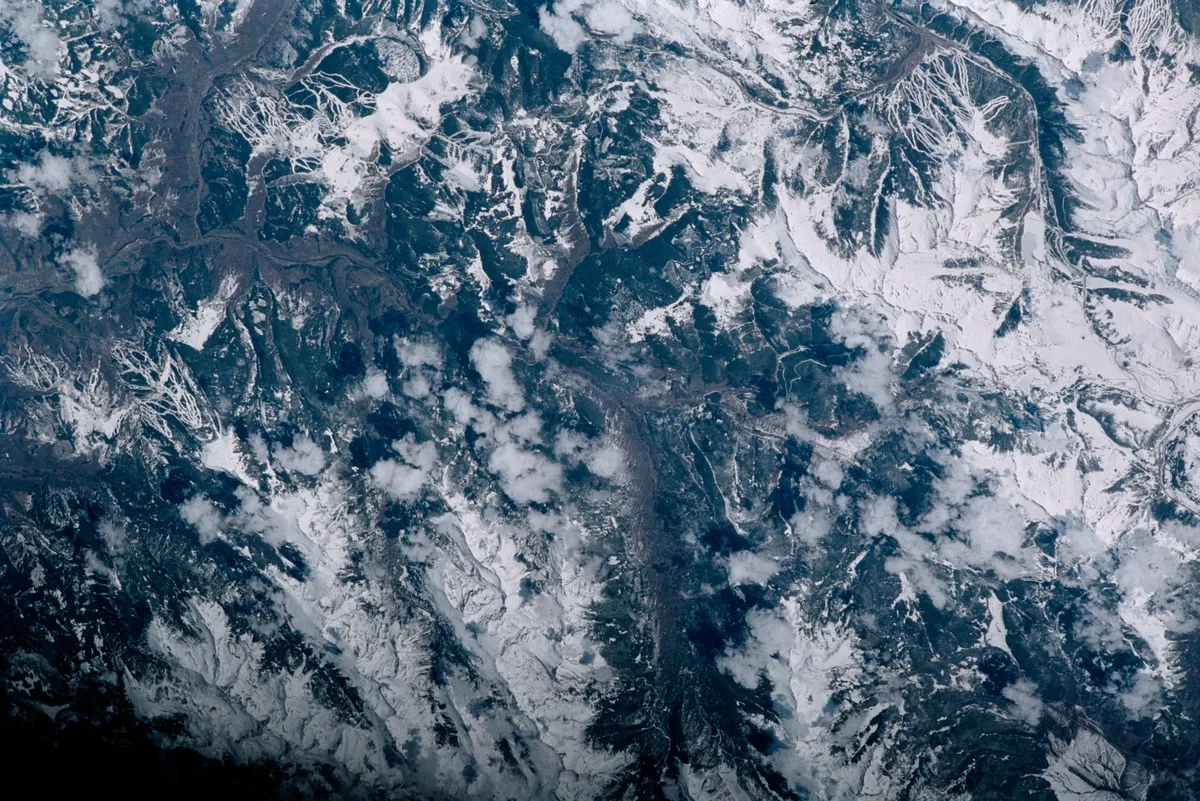 Snow-covered mountain ridges and dark valleys spread across the frame in a high-altitude aerial view, with scattered clouds casting soft shadows over the terrain. The scene has a cold blue-gray winter tone.
