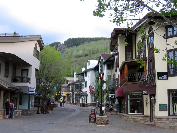 A pedestrian village street in Vail is lined with Bavarian-style alpine buildings, stone bases, and balconies, with a lamppost and shopfronts framing the curving road. Green hills rise in the background beneath a pale blue-hour sky, and a few people are walking through the quiet scene.