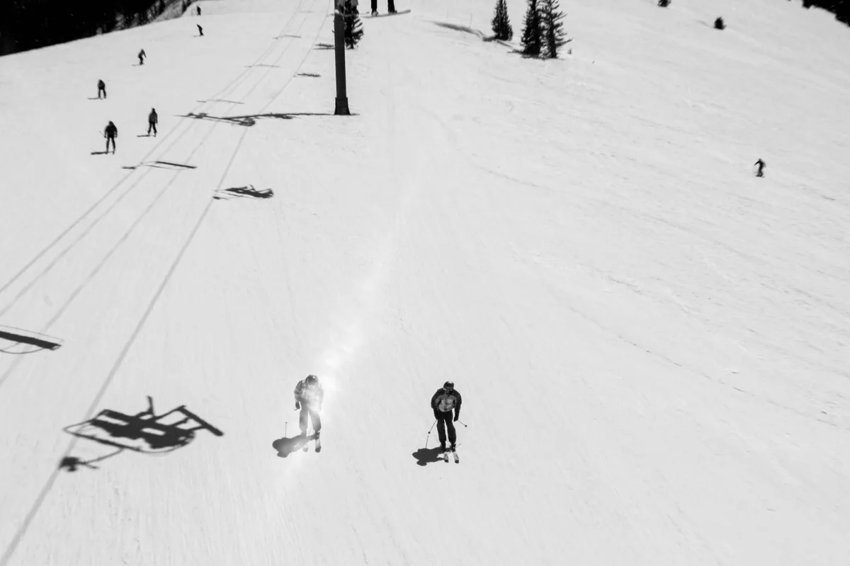 Skiers are scattered across a wide, sunlit slope of smooth white snow, with long chairlift shadows stretching diagonally across the hill. One skier catches bright glare near the center, while lift towers and a few distant riders punctuate the open mountain scene.