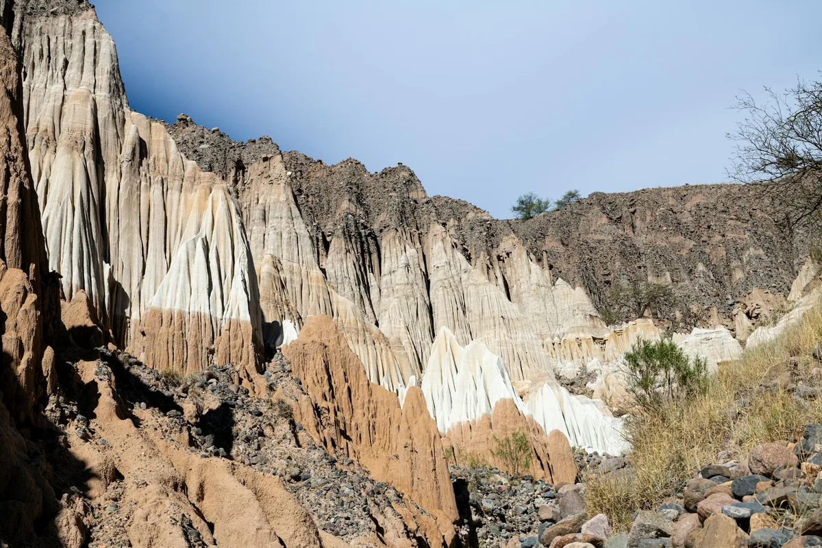 Sunlit white and pale tan volcanic tuff hoodoos rise in layered spires above a rocky slope, with dark volcanic rubble and sparse brush in the foreground. A clear blue sky and the rough canyon wall emphasize the remote, eroded landscape.