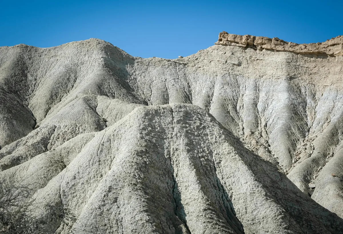Weathered pale-gray tuff formations fill the frame in steep, ribbed ridges and gullies, with a jagged tan caprock line running along the top. Bright blue sky above and sparse dark brush below emphasize the remote, eroded landscape.