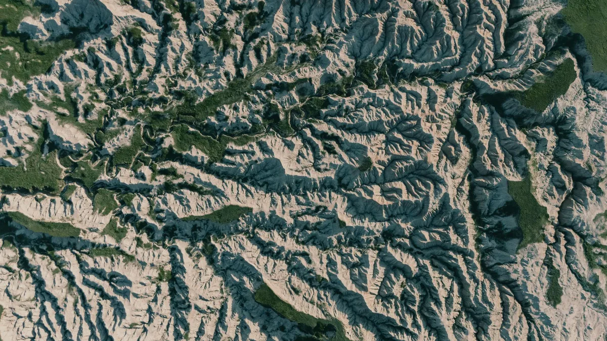 An aerial view shows Wheeler Geologic Area’s pale volcanic tuff carved into a dense maze of ridges, gullies, spires, and hoodoos, with dark green forest strips cutting through the eroded formations. Low golden-hour light emphasizes the ridged texture and layered shadows across the remote landscape.