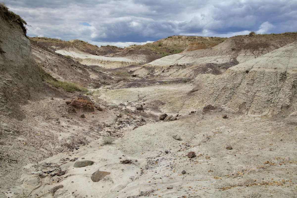 Eroded pale volcanic tuff hills and gullies stretch across the frame, with rounded ridges and scattered rocks leading into the distance. Dark, heavy clouds hang overhead, contrasting with the light cream and tan layers of the remote badlands.
