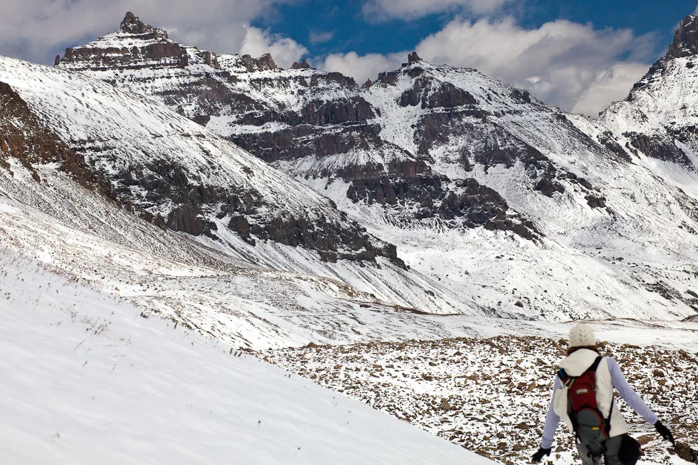 Snow blankets the steep alpine basin below jagged, dark mountain ridges, with layered cliffs and a patch of blue sky overhead. In the lower right, a hiker in a red backpack walks across the white slope, emphasizing the scale of the landscape.