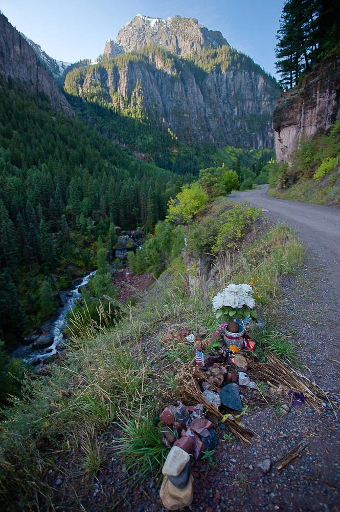 A narrow gravel road curves along a steep alpine slope above a rushing creek, with dark pine-covered canyon walls and a sheer sunlit peak rising in the distance. In the foreground, a small roadside memorial of flowers, candles, and stacked stones sits beside the trail in the morning light.